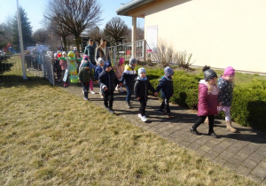 Dzieci wraz z paniami maszerują z wiosennymi plakatami, panią Wiosną pod budynkiem przedszkola.